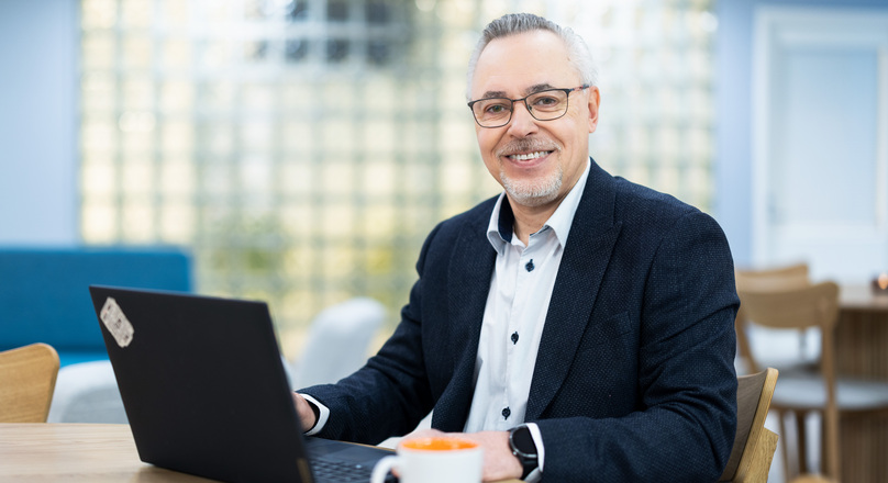 Silmälasipäinen henkilö istuu pöydän ääressä hymyillen kameralle.