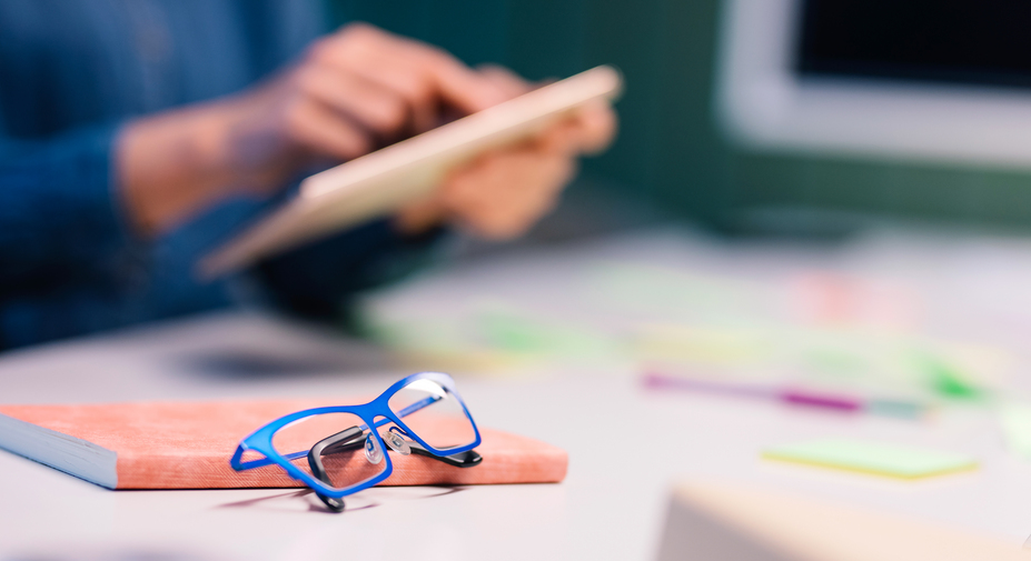 Silmälasit ja kalenteri pöydällä ja henkilö taustalla tabletti kädessä.