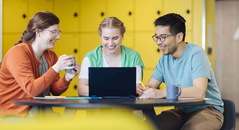 Insinööriopiskelijat tekevät pöydän ääressä ryhmätyötä. Pöydällä on kyniä, kahvikuppi ja tietokone.