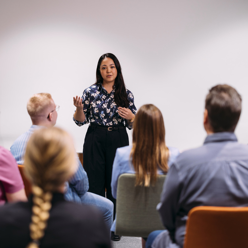 Henkilö puhuu luentosalin edessä, ihmiset istuvat tuoleilla ja kuuntelevat.