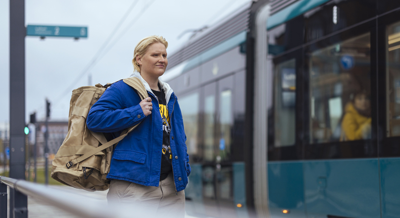 Sinipaitainen henkilö seisoo ratikkapysäkillä reppu selässä.