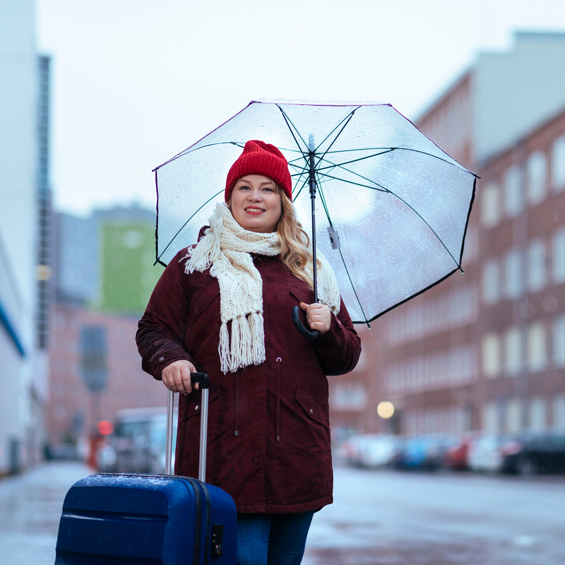 Punapipoinen henkilö seisoo ulkona matkalaukun kanssa. On sateinen syksykeli. Hänellä on läpinäkyvä sateenvarjo auki.