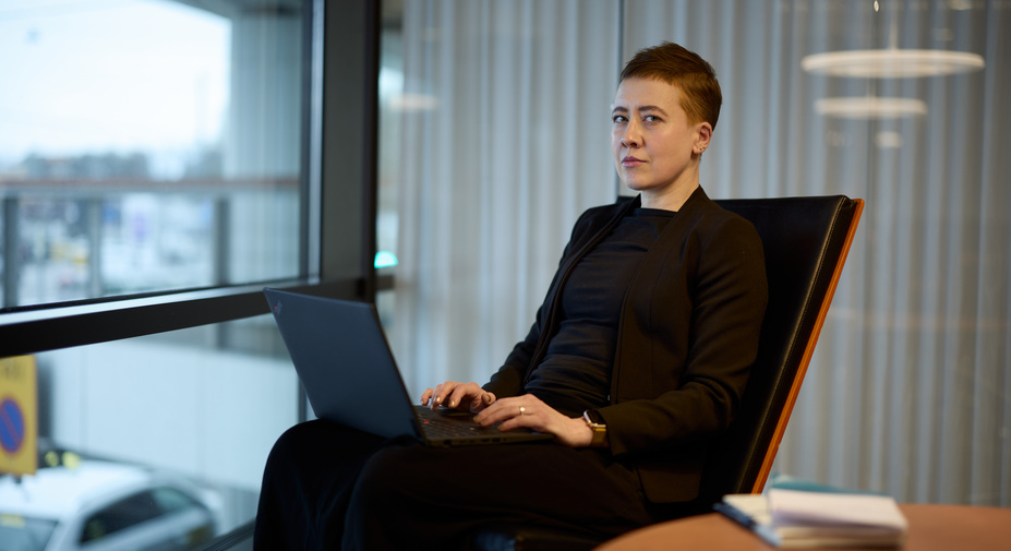 Henkilö istuu, katsoo kameraan ja pitää sylissään kannettavaa tietokonetta.