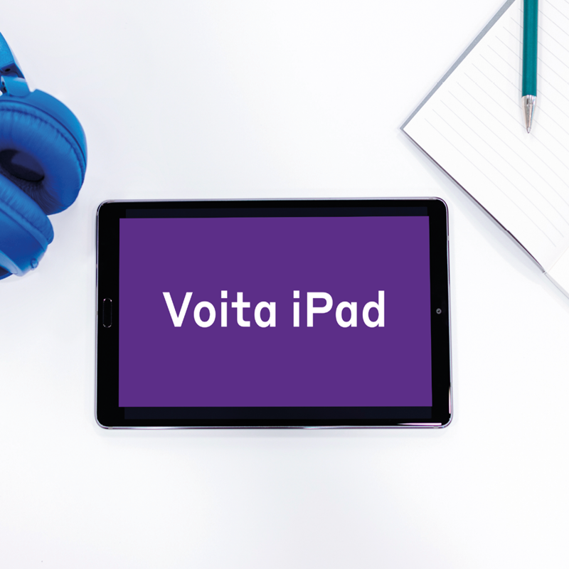 Pöydällä on tabletti, vihko, kynä ja kuulokkeet. Kuva on kuvattu suoraan ylhäältä. Näytöllä lukee voita iPad.