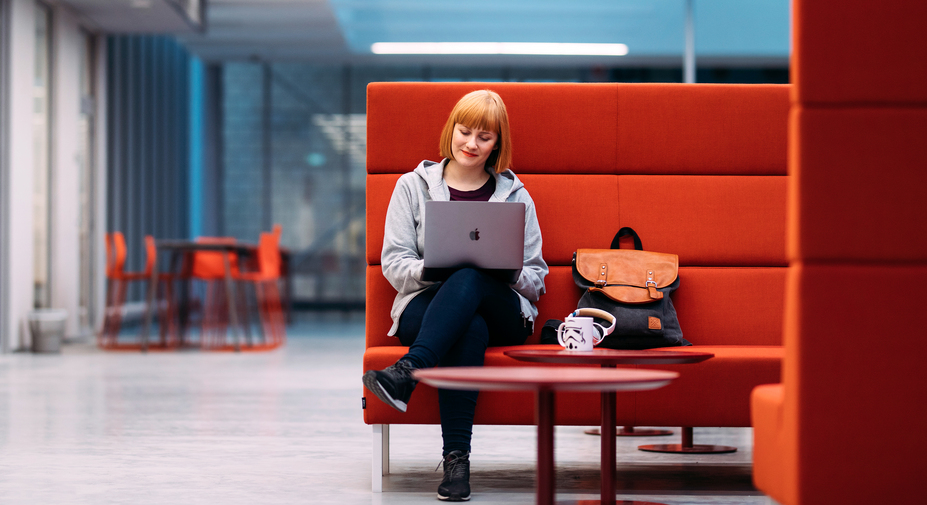 Opiskelija istuu oppilaitoksen aulassa punaisessa sohvassa tietokone sylissään.