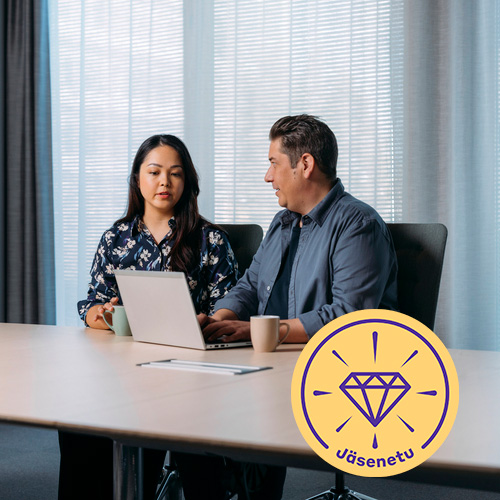 Kaksi toimistotyöntekijää istuu tietokoneen ääressä neuvotteluhuoneessa keskustelemassa. Toisen kädet ovat näppäimistöllä. Pöydällä on kahvikupit.