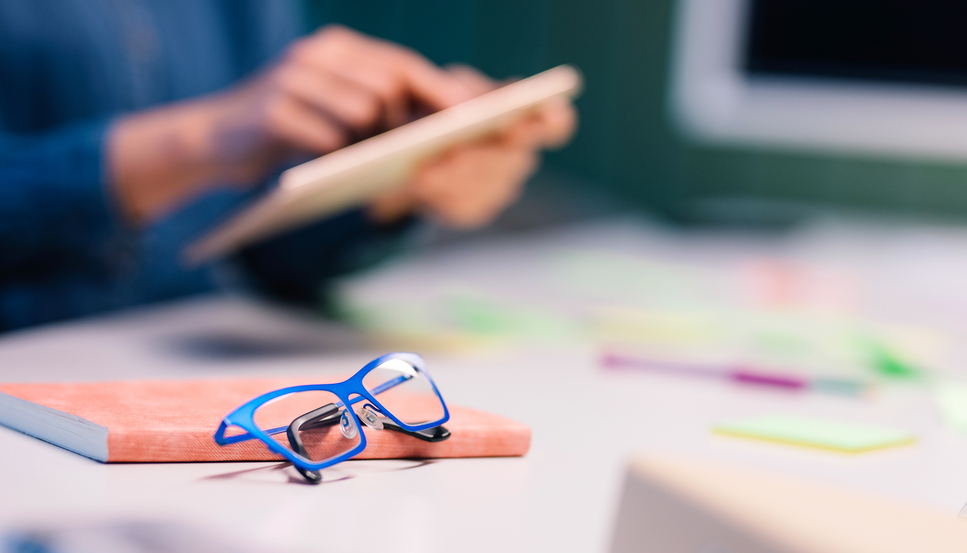 Silmälasit ja kalenteri pöydällä ja henkilö taustalla tabletti kädessä.