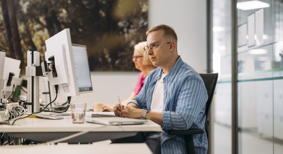 Kaksi toimistotyöntekijää työskentelee tietokoneen ääressä toimistossa vierekkäin. Toinen tekee muistiinpanoja.