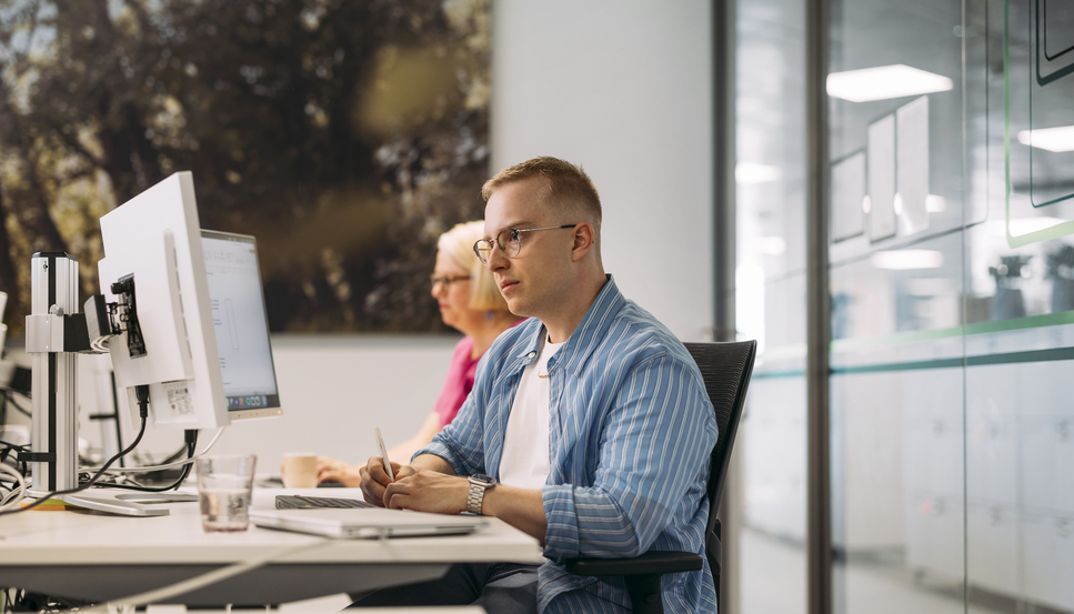 Kaksi toimistotyöntekijää työskentelee tietokoneen ääressä toimistossa vierekkäin. Toinen tekee muistiinpanoja.