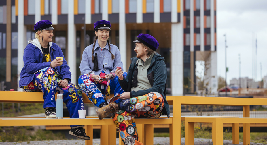 Kolme opiskelijaa juttelee pöydillä insinöörihatut päässä.