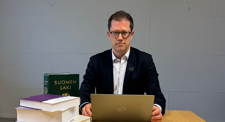 Henkilö istuu pöydän ääressä auki olevan kannettavan tietokoneen takana. Vieressä lakikirjoja.