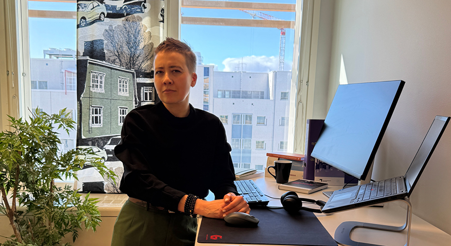 Henkilö nojaa työpöytään ja katsoo kameraan totisesti