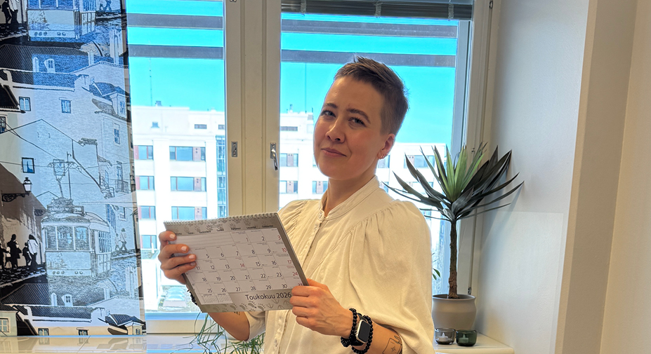 Henkilö katsoo kevyesti hymyillen kameraan pidellen seinäkalenteria käsissään