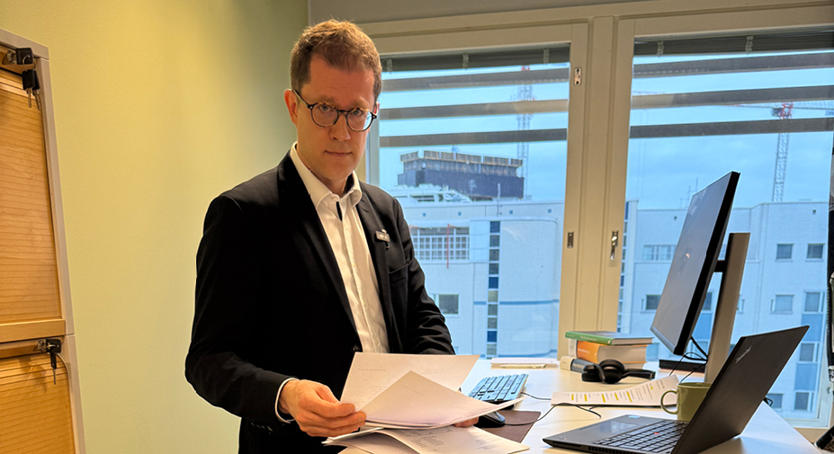 Henkilö on työpisteellään ja pitää kädessä papereita. Katsoo kameraan huolestuneena.