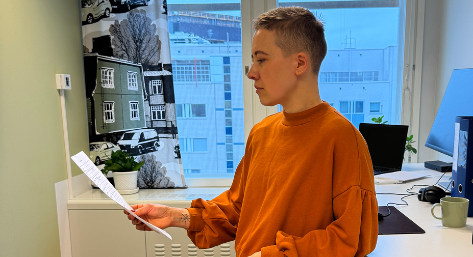 Henkilö pitää kädessään paperia ja lukee sitä työpisteellään seisten.